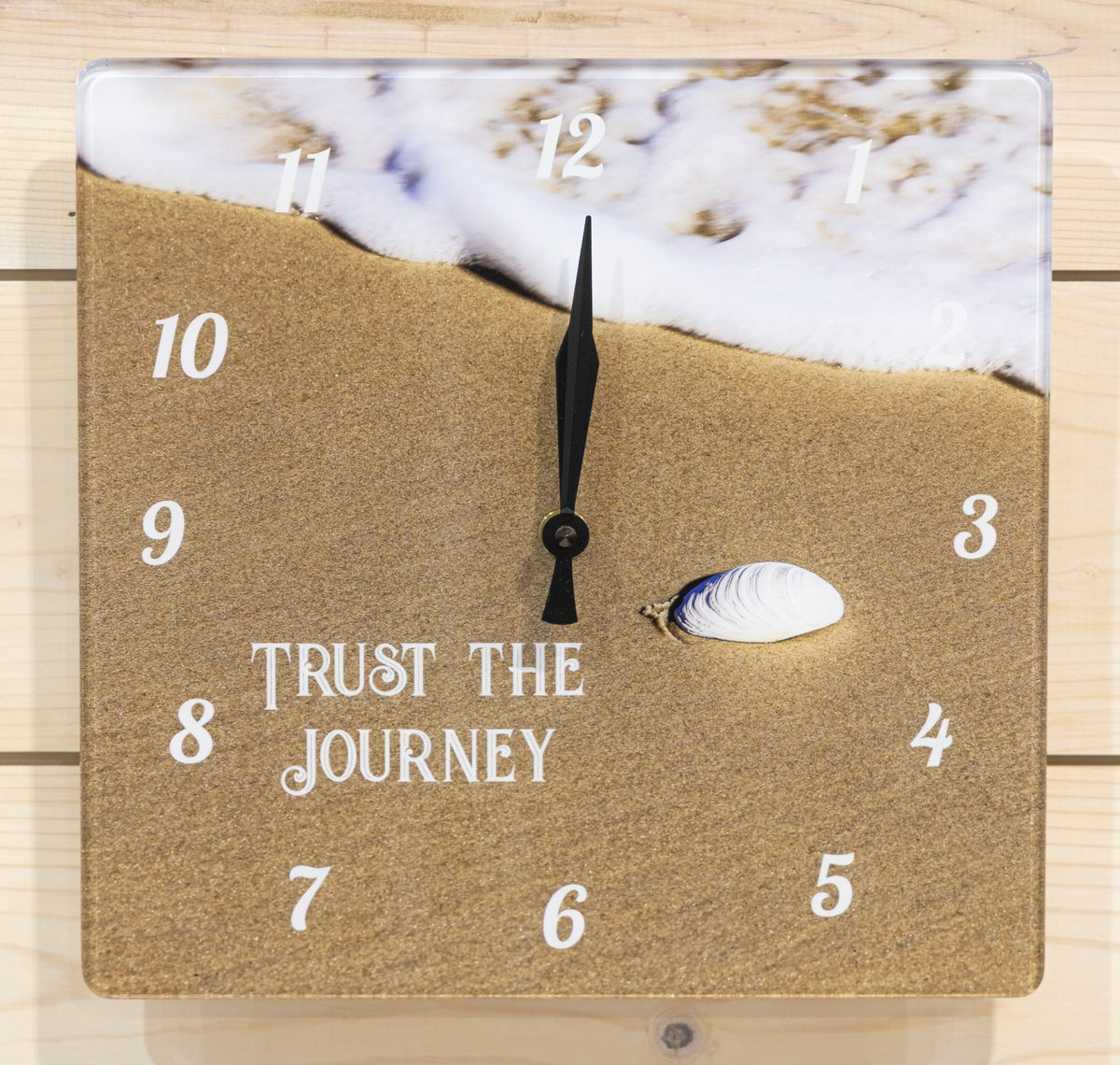 Square wall clock featuring a beach, seashell, and ocean waves. White Arabic numeral hour markers are placed over the scene, and the center reads the phrase 'Trust the Journey' in White script.